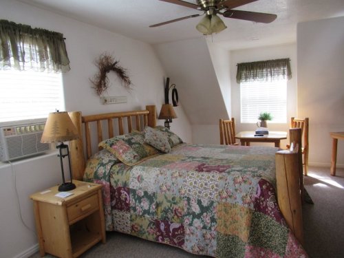Cozy bedroom with a wooden bed, colorful quilt, table, chairs, and air conditioning unit by the window.