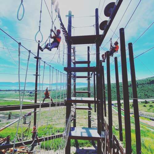 A person climbs a rope structure in an outdoor adventure park, surrounded by green hills and a clear blue sky.