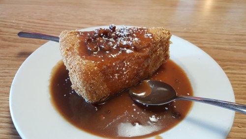 A slice of dessert topped with a glossy sauce and sprinkled with powdered sugar, served on a white plate with a spoon.