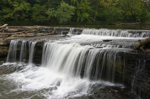 A serene waterfall cascading over rocky ledges, surrounded by lush green trees and a calm river.
