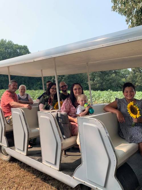 A group of seven people, including a child, sitting on a tram with sunflowers, surrounded by greenery.