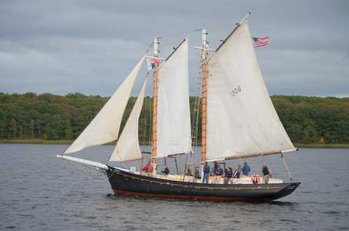 A classic sailing ship with white sails and an American flag, navigating calm waters with a crew on board.