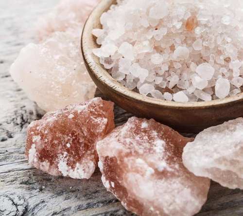 A wooden bowl filled with coarse pink salt, surrounded by larger pink salt crystals on a textured surface.