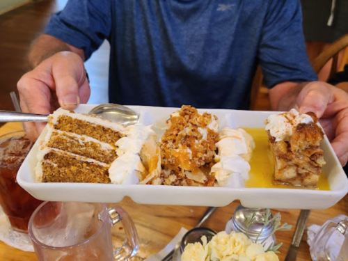 A person holds a platter with three desserts: carrot cake, a layered dessert with nuts, and a slice of apple pie with whipped cream.