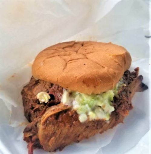 A close-up of a pulled meat sandwich with coleslaw on a soft bun, resting on white paper.