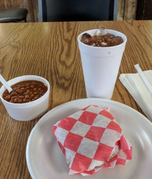 A plate with a wrapped sandwich, a cup of baked beans, and a drink in a white cup on a wooden table.