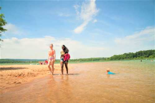 Two women walk along the shore of a beach, with a child swimming nearby and a clear blue sky above.