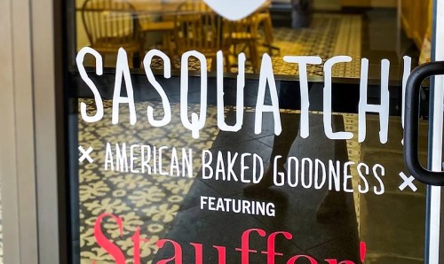 Sign on a glass door reading "Sasquatch! American Baked Goodness" featuring Stauffer's, with a decorative floor visible inside.