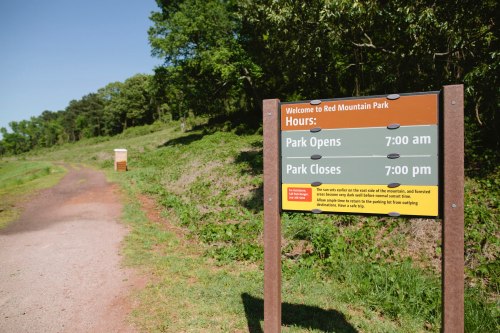 Sign at Red Mountain Park displaying hours: Opens at 7:00 AM and closes at 7:00 PM, with a scenic path in the background.