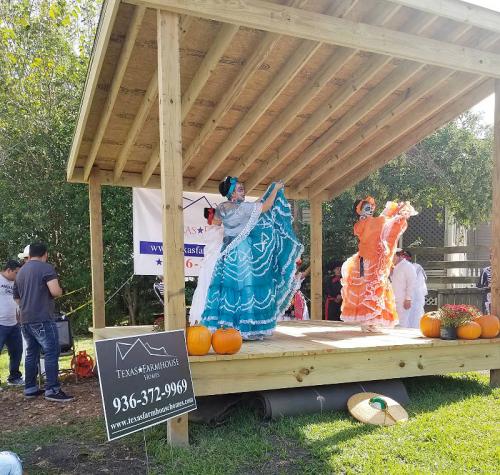 Two dancers in colorful traditional dresses perform on a wooden stage, surrounded by pumpkins and an audience.