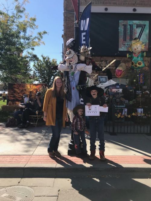 A woman and two children pose in front of a decorated storefront with a scarecrow display on a sunny day.