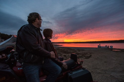 A couple watches a vibrant sunset over a lake, seated on an ATV by the shore.