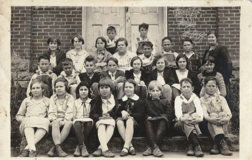 A black-and-white photo of a large group of children and adults seated on steps in front of a brick building.
