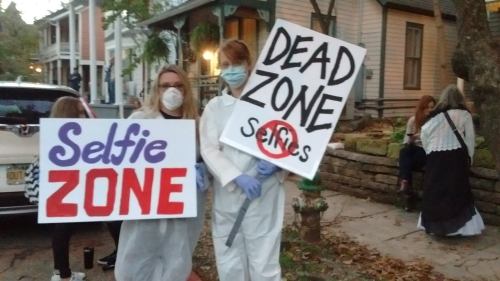 Two people hold signs reading "Selfie Zone" and "Dead Zone" while wearing masks and gloves in a festive outdoor setting.