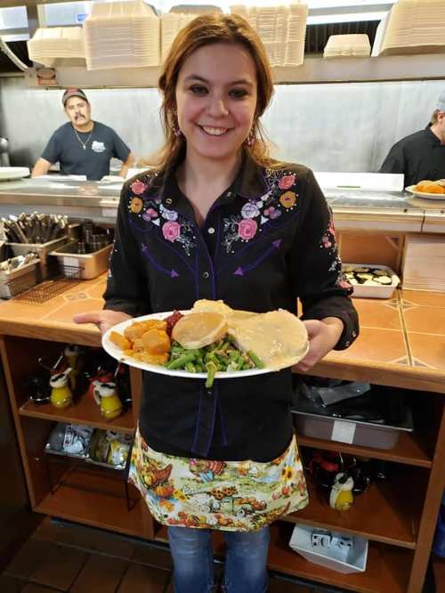 A smiling woman holds a plate of food in a restaurant, with a kitchen and staff visible in the background.