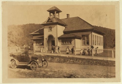 Historic school building with a bell tower, children playing outside, and an old car parked nearby.