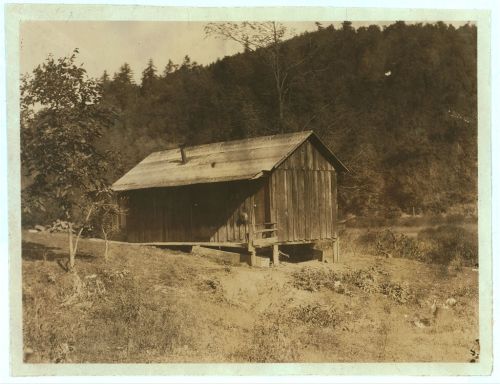 A rustic wooden cabin on stilts, surrounded by trees and a grassy area, set against a hilly backdrop.