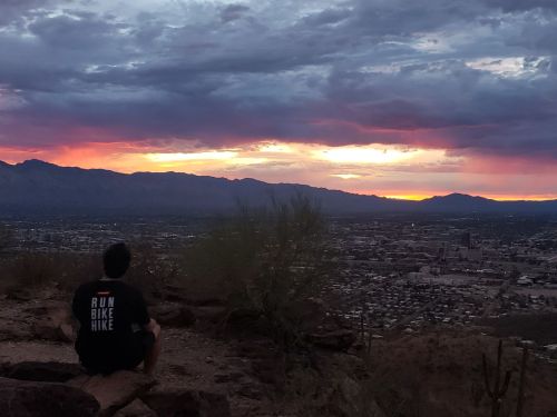 A person sits on a rocky ledge, watching a colorful sunset over a city and mountains in the background.