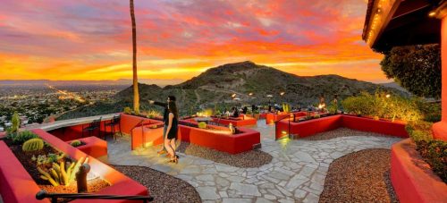 A vibrant sunset over a scenic outdoor patio with red walls, surrounded by mountains and city lights.