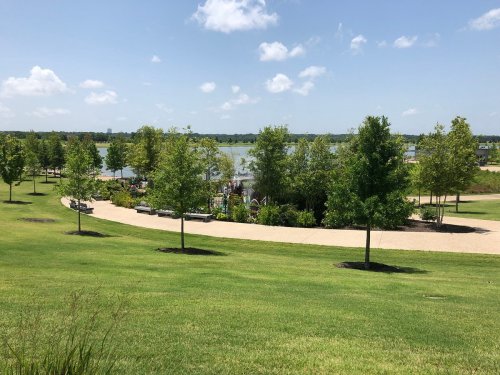 A scenic park with green lawns, trees, and a winding path leading to a lake under a clear blue sky.