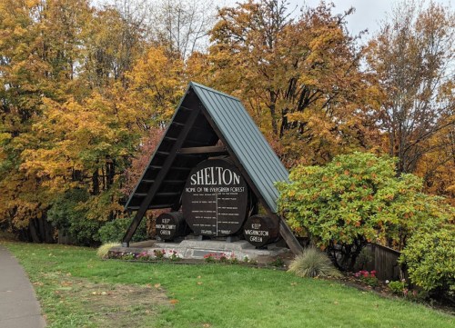 A wooden sign reading "Shelton, Home of the Evergreen Forests," surrounded by autumn foliage and greenery.
