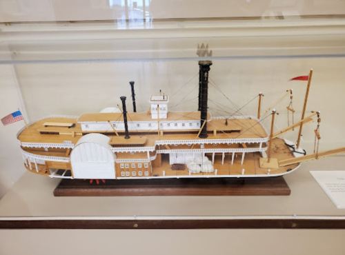 A detailed model of a steamboat displayed in a glass case, featuring multiple smokestacks and an American flag.