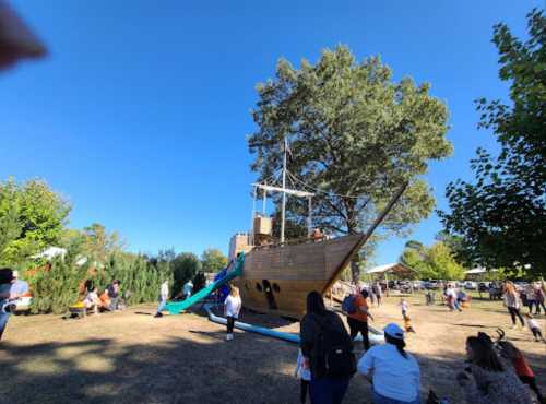 A wooden pirate ship playground with a slide, surrounded by families enjoying a sunny day in a park.