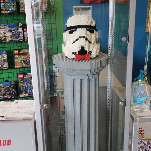 A large LEGO sculpture of a Stormtrooper's head on a column, displayed in a glass case at a store.