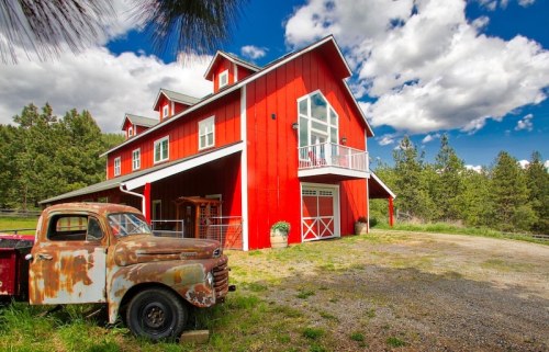 A rustic red barn-style house with a vintage truck in front, set against a backdrop of trees and a blue sky.