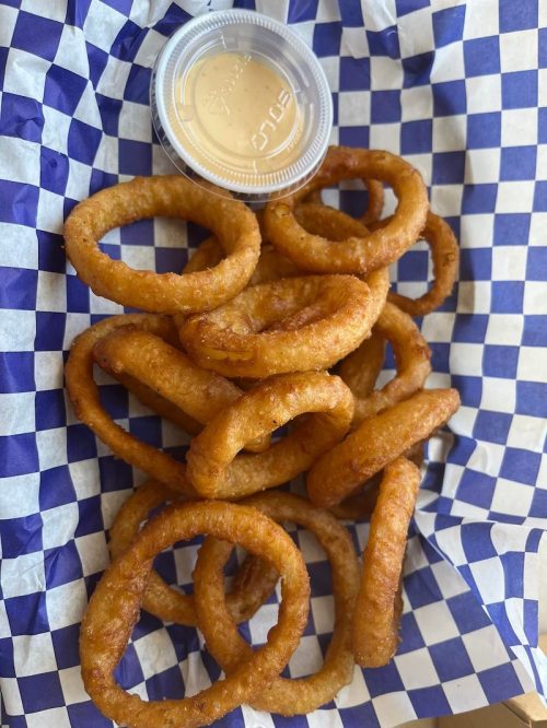 A serving of crispy onion rings arranged on a blue and white checkered paper, accompanied by a small cup of dipping sauce.