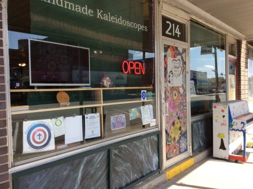 A storefront with a bright "OPEN" sign, showcasing handmade kaleidoscopes and colorful decorations in the windows.