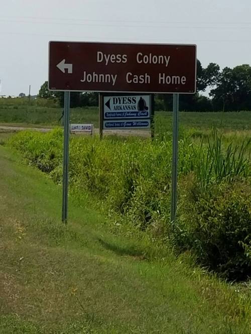 Signpost directing to Dyess Colony and Johnny Cash Home in Arkansas, surrounded by green fields.