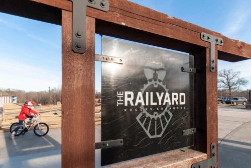 Sign for "The Railyard" in Rogers, Arkansas, with a child riding a bike in the background.
