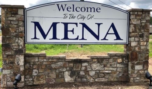 Welcome sign for the city of Mena, featuring a stone base and blue lettering against a cloudy sky.