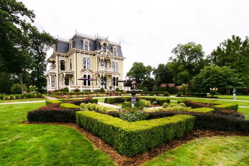 A grand Victorian mansion surrounded by manicured gardens and hedges, featuring a central fountain and lush greenery.