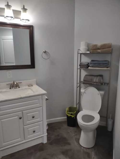 A clean bathroom featuring a toilet, sink with a mirror, and a shelf with neatly stacked towels.