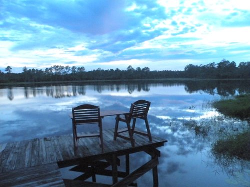 Two wooden chairs on a dock overlooking a calm lake, reflecting the blue sky and clouds at dusk.