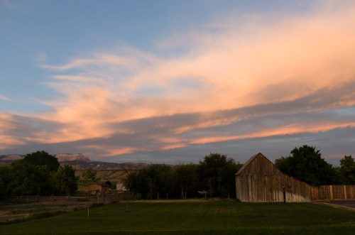 A rustic barn sits in a green field under a colorful sunset sky with soft clouds and distant mountains.
