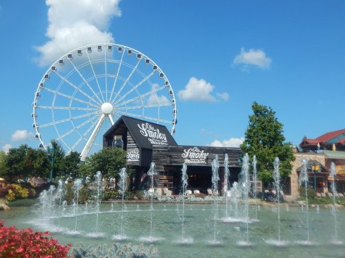 A large Ferris wheel towers over a fountain and a rustic building in a vibrant outdoor setting.