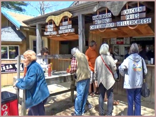 A busy outdoor food stand with people ordering, featuring a menu of burgers, fries, and drinks.