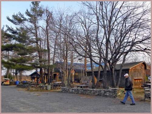 A man walks past rustic buildings surrounded by bare trees in a quiet outdoor setting.