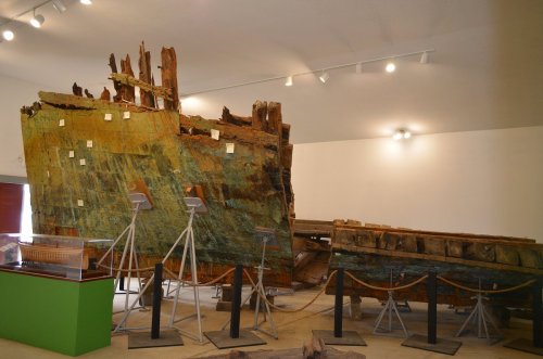 A large, partially restored shipwreck displayed in a museum, with wooden beams and artifacts on stands.