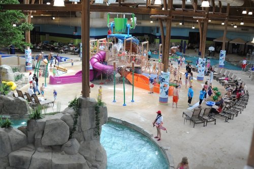 Indoor water park featuring slides, play structures, and children enjoying the water, surrounded by lounge chairs and greenery.