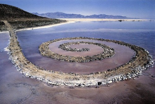 A spiral stone formation on a dry lakebed, surrounded by water and mountains in the background.