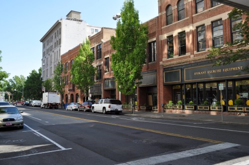 Street view of a city block featuring brick buildings, trees, parked cars, and a storefront for Spokane Exercise Equipment.