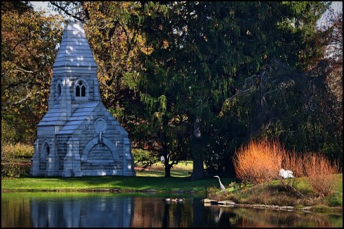 A stone chapel beside a calm pond, surrounded by trees and autumn foliage, with a heron near the water's edge.