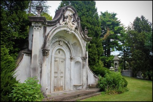 A weathered mausoleum with ornate architecture, surrounded by lush greenery in a serene cemetery setting.