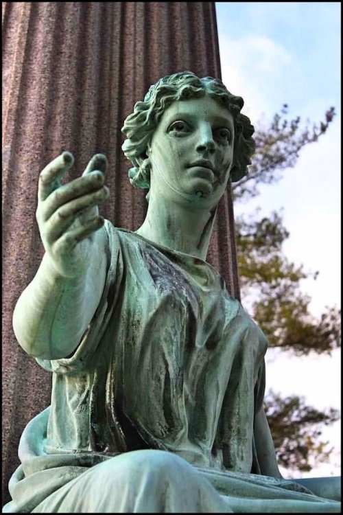 A close-up of a weathered bronze statue of a woman, seated with an outstretched hand, against a backdrop of trees.
