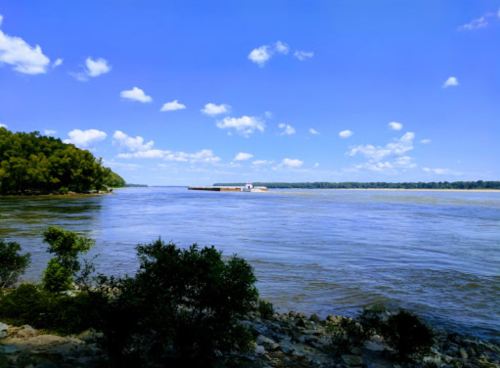 A serene river scene with blue skies, fluffy clouds, and a barge in the distance, surrounded by lush greenery.
