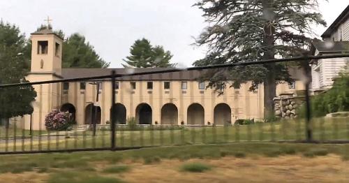 A beige building with arches and a bell tower, surrounded by trees and a fence, viewed from a moving vehicle.